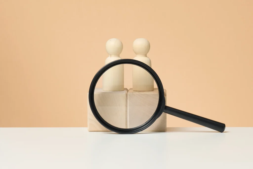 Suurennuslasi kohdistettuna puufiguureihin symboloimassa saavutettavuustestausta, yksityiskohtien tarkastelua ja käyttäjien huomioimista digitaalisissa palveluissa.