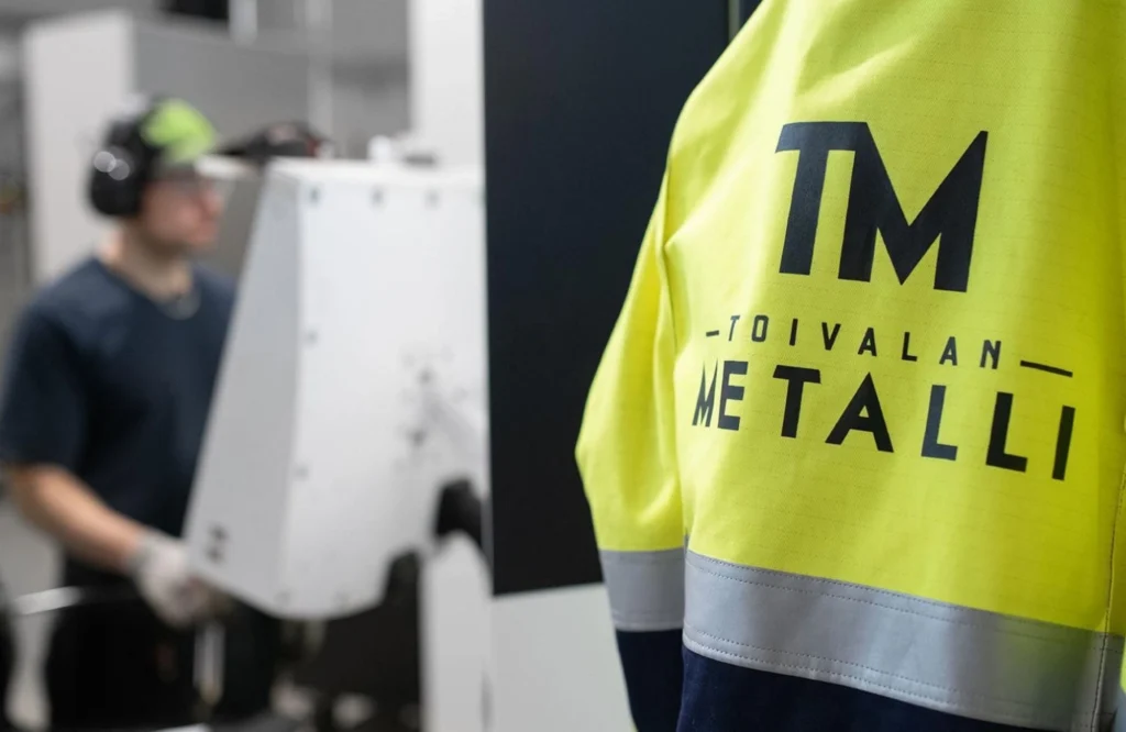 Toivalan Metallin työntekijä huomiovaatteessa, jossa näkyy yrityksen logo. Taustalla työntekijä työskentelee metalliteollisuuden tuotantotiloissa.